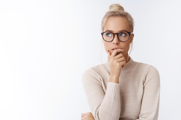 Blogger de estilo de vida femenino creativo con cabello rubio peinado en moño con gafas y un suéter tocando el labio pensativo mirando la esquina superior izquierda, pensando en hacer un plan