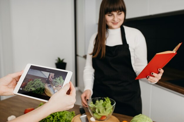 Blogger femenina grabando a sí misma mientras prepara la comida y lee un libro