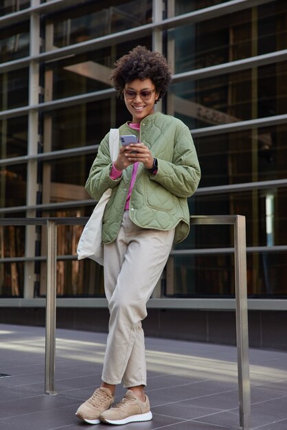 Blogger femenina positiva de moda sostiene que el teléfono móvil usa tecnología moderna para llamar en línea y las redes sociales usan gafas de sol de moda, un atuendo elegante se encuentra afuera envía sms a un amigo