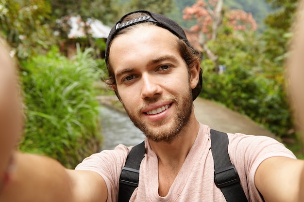 Blogger masculino barbudo joven con estilo con mochila posando al aire libre mientras graba videos o toma selfie