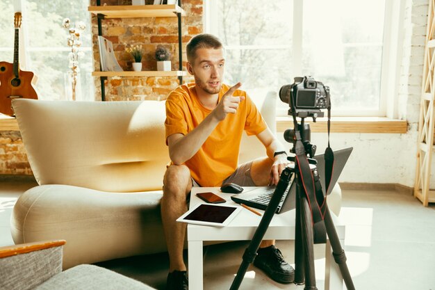 Blogger masculino caucásico joven con revisión de video de grabación de cámara profesional de gadgets en casa. Blogs, videoblogs, vlogs. Hombre haciendo vlog o transmisión en vivo sobre fotografía o novedad técnica.