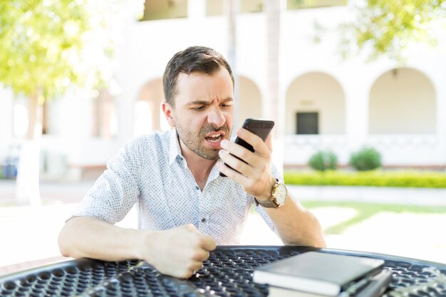 Blogger masculino furioso gritando en el teléfono móvil mientras está sentado en la mesa en el jardín