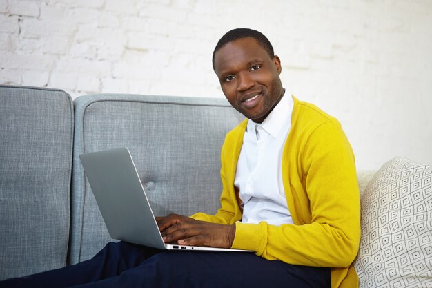 Blogger masculino guapo de piel oscura en cardigan amarillo mecanografía en una computadora portátil genérica, publicando una nueva publicación en su blog en línea, habiendo inspirado expresión, mirando y sonriendo