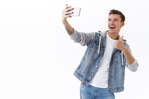 Foto gratuita blogger masculino guapo tomando selfie en teléfono inteligente, muestra el pulgar hacia arriba en señal de aprobación, recomienda un buen lugar a los seguidores