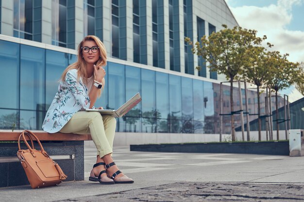 Blogger de moda elegante relajándose al aire libre, trabajando en la computadora portátil, sentado en un banco contra un rascacielos.