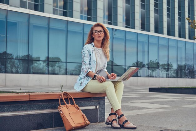 Blogger de moda elegante relajándose al aire libre, trabajando en la computadora portátil, sentado en un banco contra un rascacielos.