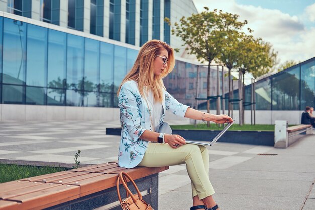 Blogger de moda elegante relajándose al aire libre, trabajando en la computadora portátil, sentado en un banco contra un rascacielos.