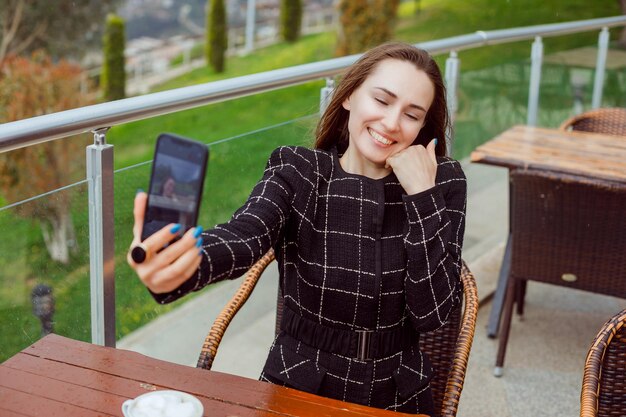 El blogger sonriente se está tomando una selfie con la cámara trasera del teléfono inteligente sosteniendo la mano en la mejilla en el fondo de la vista de la naturaleza