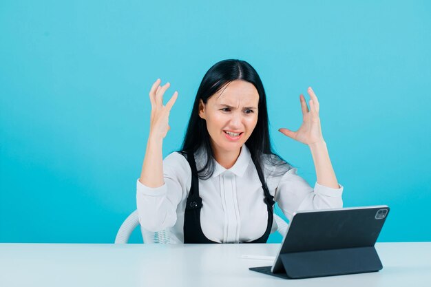 La bloguera enojada está levantando las manos mirando la cámara de la tableta con fondo azul