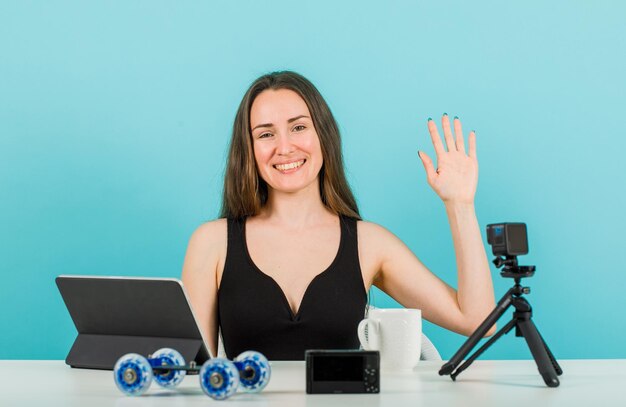 Una bloguera sonriente está mirando a la cámara mostrando un gesto de hola con fondo azul