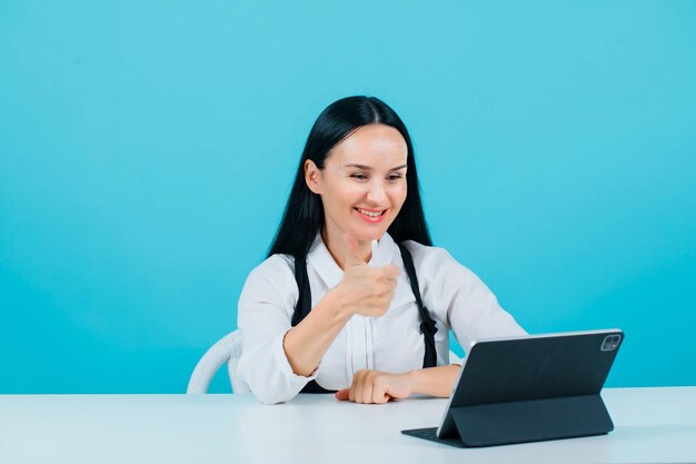 La bloguera sonriente está mostrando un gesto perfecto al mirar la cámara de su tableta con fondo azul