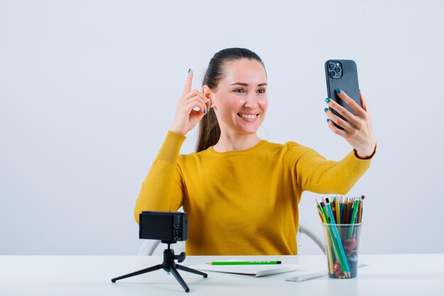 Una bloguera sonriente se está tomando una selfie señalando con el dedo índice sobre fondo blanco