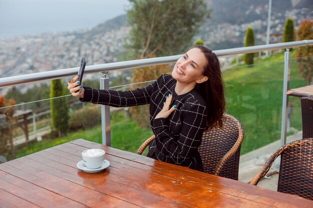 Una bloguera sonriente se está tomando una selfie con su teléfono inteligente mirando hacia arriba sentada en el fondo de la vista de la ciudad