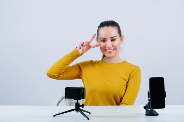 Una bloguera sonriente posa mostrando dos gestos frente a su mini cámara con fondo blanco