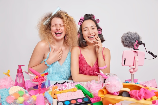 Las blogueras de belleza femeninas llenas de alegría graban video ríen felizmente se divierten juntos usan un vestido y hacen un peinado para ocasiones especiales rodeadas de diferentes productos cosméticos aislados sobre fondo blanco