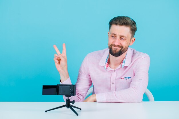 Un bloguero sonriente posa para su mini cámara mostrando dos gestos en el fondo azul