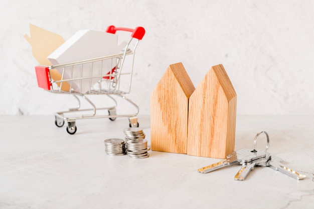 Bloques de casas de madera; pila de monedas; Llaves y pequeño carrito de compra sobre fondo de hormigón.