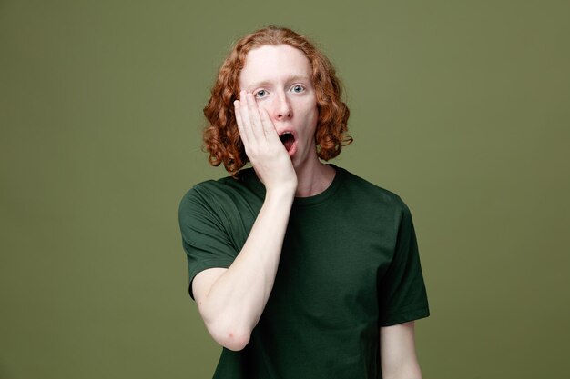 Boca cubierta sorprendida con la mano joven guapo con camiseta verde aislado sobre fondo verde