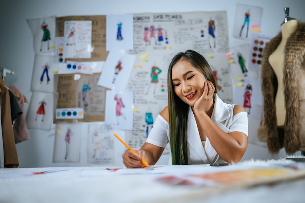 Boceto de moda de dibujo femenino joven diseñador asiático en taller. Hermosa ropa dibujo imagen a bordo detrás de ella