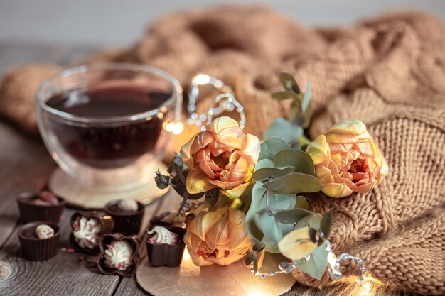 Bodegón festivo con una bebida en una taza, chocolates y flores sobre un fondo borroso.