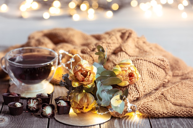 Bodegón festivo con una bebida en una taza, chocolates y flores sobre una mesa borrosa con bokeh.