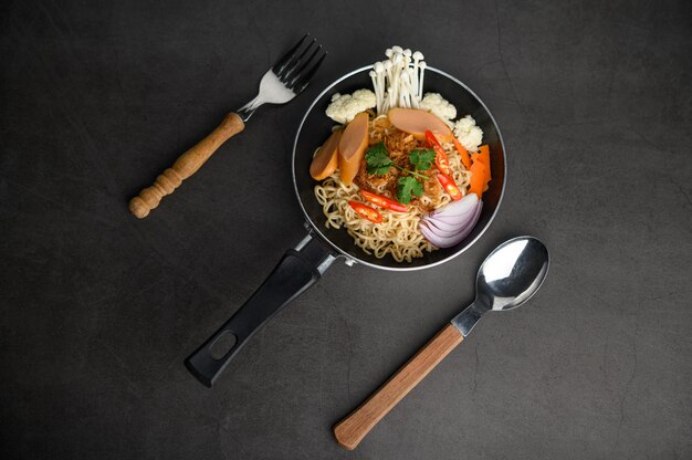 Bodegón con los fideos en una sartén, tenedor y cuchara en el piso de cemento negro.