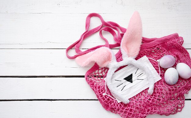 Bodegón de Pascua con bolsa de hilo rosa, orejas de conejo de Pascua decorativas, máscara médica y huevos sobre una superficie de madera.