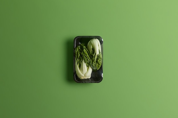 Bok choy o repollo chino envuelto con film alimenticio en bandeja negra. Hortalizas frescas para la venta en el supermercado aislado sobre fondo verde. Concepto de estilo de vida saludable, refrescos y nutrición.