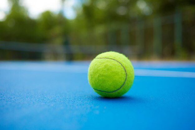 La bola amarilla está tendida en la alfombra azul de la cancha de tenis. Plantas verdes borrosas y árboles detrás.