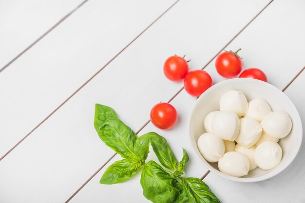 Bola de queso mozzarella con hojas de albahaca y tomates rojos sobre fondo blanco de madera