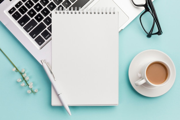 Bolígrafo y cuaderno de espiral en la computadora portátil con lentes, ramita de flores y taza de café en el escritorio de oficina azul