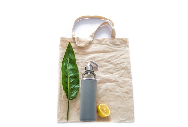 Bolsa de algodón con botella de agua frutas limones crudos hoja verde sobre pared blanca plana laical. Materiales ecológicos reutilizables sin desperdicio sin plástico