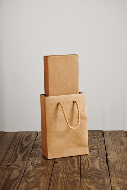 Bolsa de papel artesanal con caja de cartón en blanco en el interior presentado en mesa de madera rústica, aislado sobre fondo blanco.