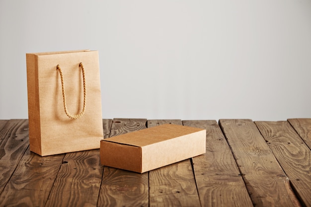 Bolsa de papel artesanal sin etiqueta junto a una caja de cartón en blanco presentada en una mesa de madera rústica, aislada sobre fondo blanco.