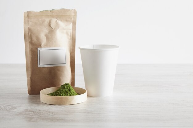 Bolsa de papel marrón artesanal, vidrio para llevar y polvo de té matcha orgánico de primera calidad en una caja sobre una mesa de madera blanca aislada sobre un fondo simple. Listo para preparar, presentación de venta.