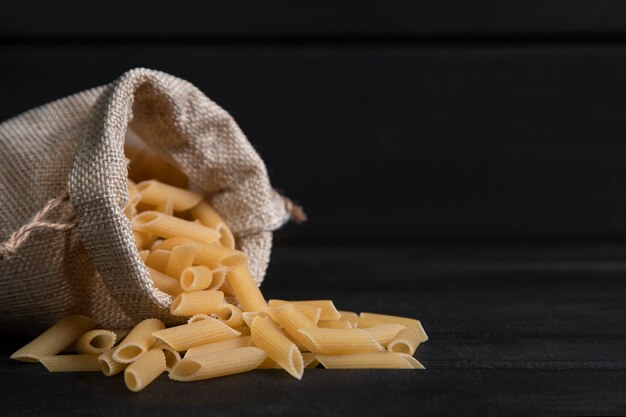 Una bolsa de saco llena de pasta penne cruda colocada sobre una mesa de madera oscura. Foto de alta calidad