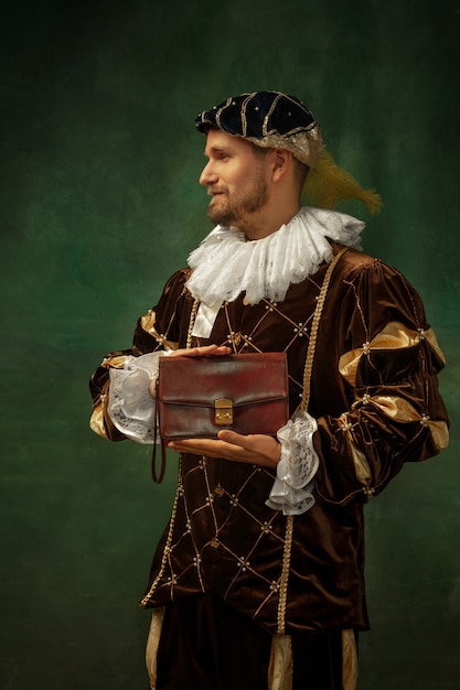 Bolso de hombre de negocios. Retrato de joven medieval en ropa vintage de pie sobre fondo oscuro. Modelo masculino como duque, príncipe, persona de la realeza. Concepto de comparación de épocas, moderno, moda.