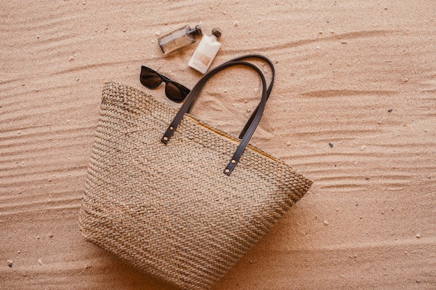 Bolso de mujer junto al perfume y gafas de sol puestas sobre arena.