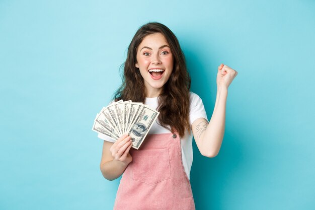 Bomba de puño de niña sonriente emocionada y mantener el premio en efectivo, ganar dinero en efectivo, recibir ingresos de algo, de pie feliz contra el fondo azul.