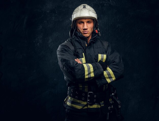 Bombero brutal en uniforme posando para la cámara de pie con los brazos cruzados y mirada confiada. Foto de estudio contra una pared de textura oscura