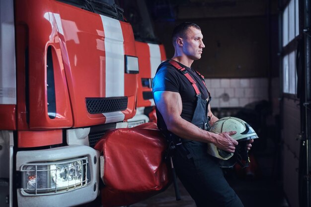 Bombero musculoso sosteniendo un casco protector en un garaje de un departamento de bomberos, apoyándose en un camión de bomberos y mirando hacia afuera