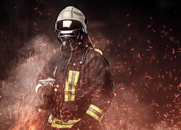 Un bombero profesional vestido con uniforme y una máscara de oxígeno parado en chispas de fuego y humo sobre un fondo oscuro.