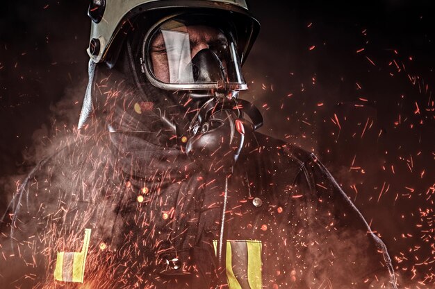 Un bombero profesional vestido con uniforme y una máscara de oxígeno parado en chispas de fuego y humo sobre un fondo oscuro.