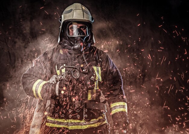Un bombero profesional vestido con uniforme y una máscara de oxígeno parado en chispas de fuego y humo sobre un fondo oscuro.