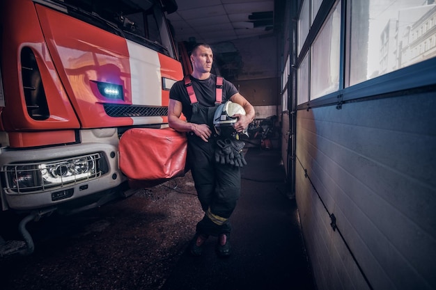 Bombero sosteniendo una chaqueta y un casco protector en un garaje de un departamento de bomberos, apoyándose en un camión de bomberos y mirando hacia afuera