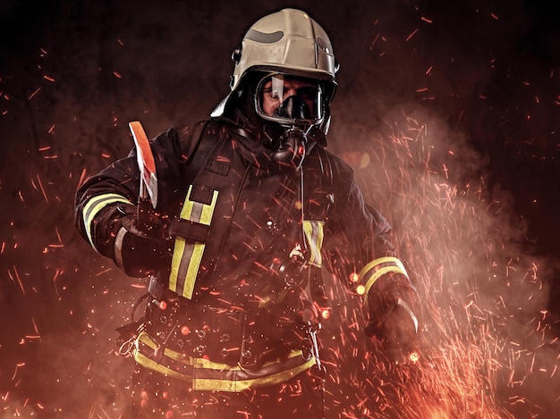 Un bombero vestido con uniforme y una máscara de oxígeno sostiene un hacha roja en chispas de fuego y humo sobre un fondo oscuro.