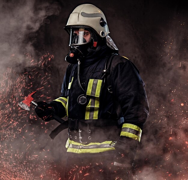 Un bombero vestido con uniforme y una máscara de oxígeno sostiene un hacha roja parada en chispas de fuego y humo sobre un fondo oscuro.