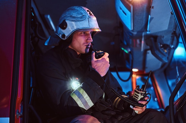 Los bomberos llegaron en horas de la noche. Bombero con uniforme protector sentado en el camión de bomberos y hablando por radio