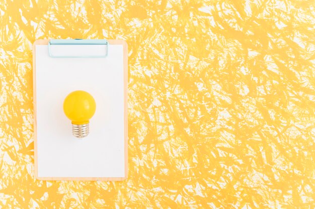 Bombilla de luz amarilla sobre papel blanco sobre portapapeles contra el fondo amarillo