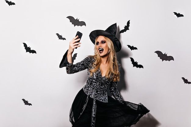 Bonita bruja con chaqueta brillante divirtiéndose en halloween. Riendo mago malvado usando el teléfono para selfie.